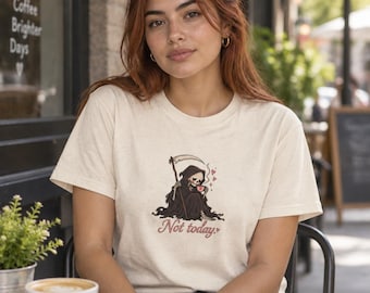 Puede incluir: Una camiseta de manga corta color crema con un gráfico de la parca sosteniendo una guadaña y una taza de café humeante. El texto "Not today." está impreso en una fuente cursiva roja debajo de la ilustración, acompañado de pequeños corazones rojos.