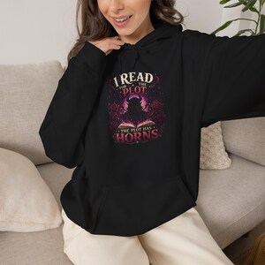 Op de afbeelding: Een zwarte hoodie met een grafische print op de borst. Het ontwerp toont een gehoornd silhouet dat uit een open boek komt, omringd door rode rozen en paars licht, met de tekst I READ FOR THE PLOT THE PLOT HAS HORNS.