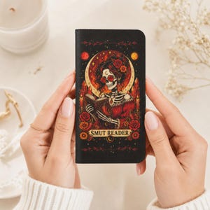 May include: A black rectangular phone case featuring a graphic illustration of a skeleton with dark hair, red glasses, and a red dress reading a book. The design includes red roses, crystals, and the text SMUT READER in a gold banner at the bottom.
