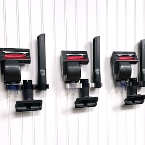 Puede incluir: Tres accesorios negros para aspiradora con cabezales de cepillo rojos están montados en una pared blanca. Cada accesorio está asegurado por un soporte de plástico transparente. Los accesorios incluyen un cabezal de cepillo, una boquilla para rincones y un cepillo pequeño.