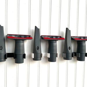 Peut inclure: Ensemble d'accessoires pour aspirateur, gris et noirs, fixés sur un mur blanc. Les accessoires comprennent différentes formes de buses, avec des accents rouges sur les têtes de brosse. L'ensemble est conçu pour le nettoyage.