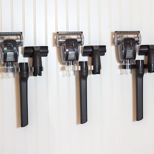 Puede incluir: Tres accesorios de aspiradora negros con contenedores de plástico transparente están colgados en una pared blanca. Los accesorios son para tapicería, grietas y limpieza de polvo.