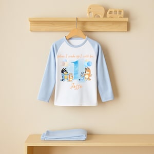 May include: A light blue and white long-sleeved pajama top featuring cartoon characters and the number 1. The text "When I wake up I will be..." and the name "Jesse" are printed on the shirt. A pair of light blue pajamas are folded below.