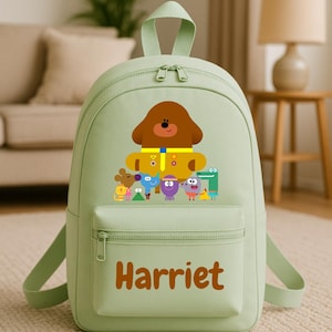 May include: A pale green backpack with a cartoon dog and other characters printed on the front. The name "Harriet" is printed in brown on the front pocket. The backpack has a top handle and adjustable straps. It is suitable for carrying school supplies.
