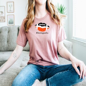 Op de afbeelding: Een roze T-shirt met een zwart-witte voetbalafbeelding in de vorm van een lip. De tekst "Bengals" staat onder de afbeelding.