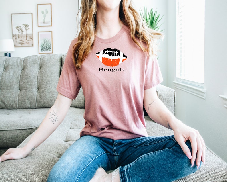 Op de afbeelding: Een roze T-shirt met een zwart-witte voetbalafbeelding in de vorm van een lip. De tekst "Bengals" staat onder de afbeelding.