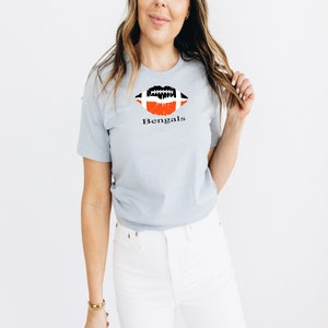 Op de afbeelding: Een lichtblauw t-shirt met een zwart en oranje voetbalvormig lipontwerp en het woord "Bengals" gedrukt op de voorkant.