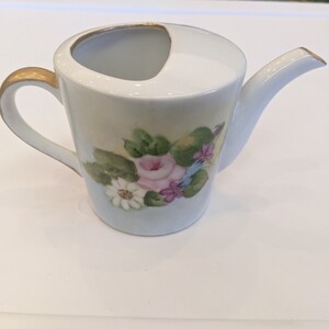 May include: A white porcelain watering can with a gold rim and a floral design. The design features pink, white, and purple flowers with green leaves.