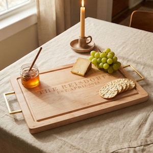 May include: A wooden serving board with gold handles, engraved with "THE REISDORFS EST. APRIL 2024". The board holds crackers, cheese, and grapes. A jar of honey with a dipper and a lit candle in a holder are also on the table.