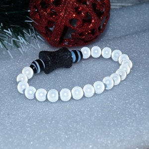 May include: A bracelet with white, circular beads and a central black, textured bead. The bracelet also includes black and white striped beads and a black bead with blue accents. The bracelet is displayed on a white, textured surface.