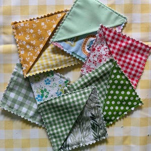 May include: A collection of triangular fabric bookmarks with pinked edges, featuring various patterns including yellow floral, light green, red and white gingham, green polka dots, blue floral, and green leaf prints, arranged in a scattered overlapping pile.