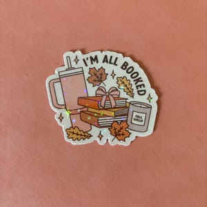 May include: A white sticker with a pink and brown illustration of a stack of books, a cup of coffee, a candle, and fall leaves. The text "I'm All Booked" is written in a circle around the illustration.