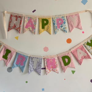 May include: A fabric bunting banner spelling out HAPPY BIRTHDAY with two rows of pennants. Each pennant features a unique floral, gingham, or patterned fabric in various colors, with contrasting felt letters stitched onto the center of each piece.
