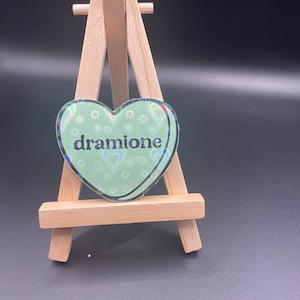 May include: A green heart-shaped sticker with a holographic finish and the text "dramione" in black lettering. The sticker is displayed on a small wooden easel.