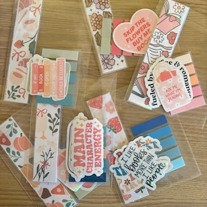 May include: A collection of colorful bookmarks and sticky notes with various designs and text. The bookmarks feature floral patterns, strawberries, and coffee cups. Text includes "Skip the flowers buy me book" and "Main Character Energy."