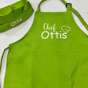 Op de afbeelding: Een groene kokschort met een witte zak en de tekst "Chef Ottis" in wit. Een bijpassende groene koksmuts met de tekst "Ottis" in wit is ook inbegrepen.