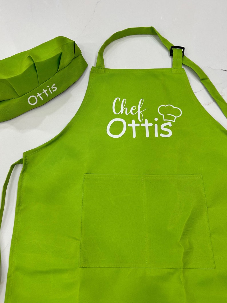Op de afbeelding: Een groene kokschort met een witte zak en de tekst "Chef Ottis" in wit. Een bijpassende groene koksmuts met de tekst "Ottis" in wit is ook inbegrepen.