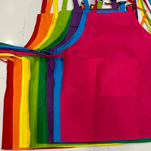 Op de afbeelding: Een set van acht kleurrijke schorten met verstelbare banden. De schorten zijn gerangschikt in een regenboogpatroon, met rood, oranje, geel, groen, blauw, indigo en violet. Elke schort heeft een grote zak aan de voorkant.