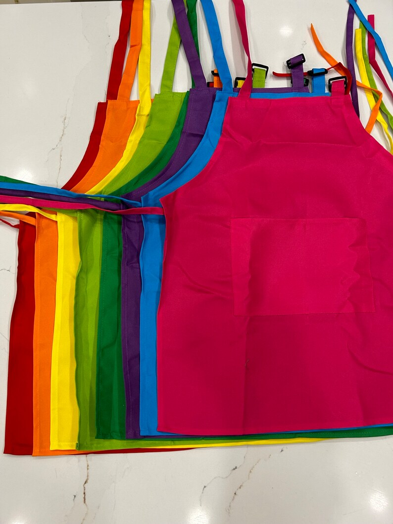 Op de afbeelding: Een set van acht kleurrijke schorten met verstelbare banden. De schorten zijn gerangschikt in een regenboogpatroon, met rood, oranje, geel, groen, blauw, indigo en violet. Elke schort heeft een grote zak aan de voorkant.