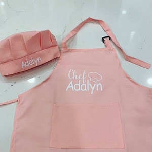 Op de afbeelding: Roze kokschort en -muts set met de naam "Addalyn" geborduurd op beide delen. De schort heeft een zak en de muts heeft een koksmuts-afbeelding.