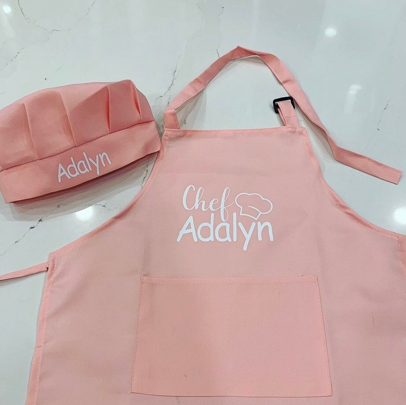 Op de afbeelding: Roze kokschort en -muts set met de naam "Addalyn" geborduurd op beide delen. De schort heeft een zak en de muts heeft een koksmuts-afbeelding.