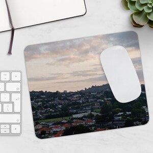 May include: A mouse pad with a scenic image of a city skyline at sunset. The sky is a soft pink and orange with fluffy white clouds. The city is in the distance and appears to be nestled in a valley.