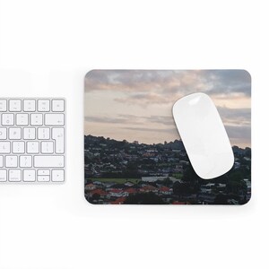 May include: A mousepad featuring a panoramic view of a city skyline at sunset. The city is nestled between hills and the sky is a soft pink and orange.