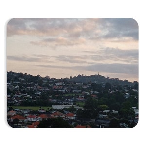 May include: A panoramic view of a suburban neighborhood with houses nestled among trees. The sky is a soft pink and gray with wispy clouds. A tall structure is visible in the distance on a hilltop.