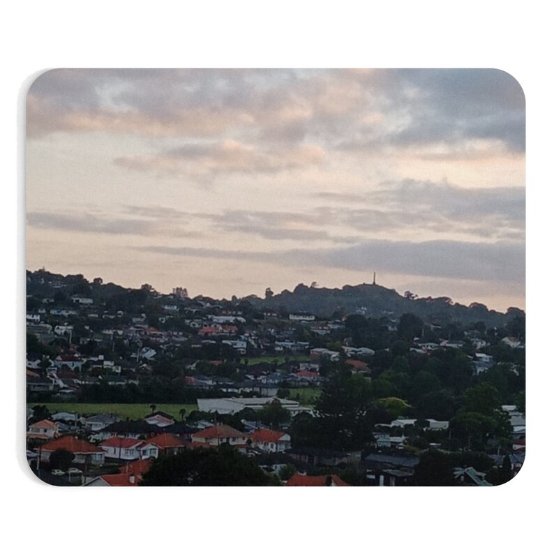 May include: A panoramic view of a suburban neighborhood with houses nestled among trees. The sky is a soft pink and gray with wispy clouds. A tall structure is visible in the distance on a hilltop.