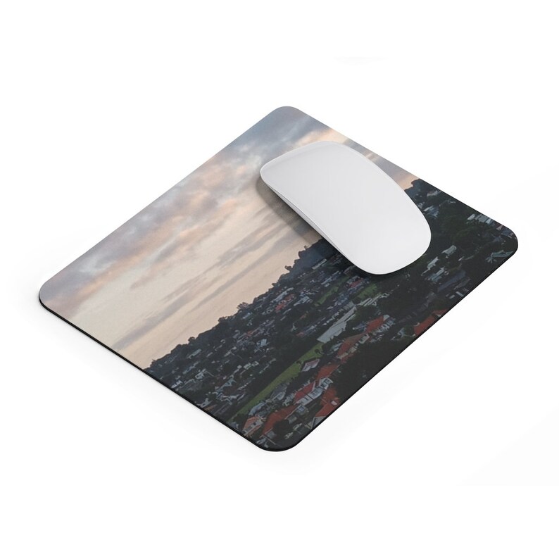 May include: A mousepad with a panoramic view of a city skyline at sunset. The city is nestled in a valley with green trees and houses. The sky is a mix of pink, orange, and purple.