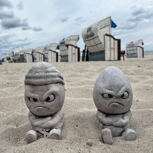 Könnte beinhalten: Zwei graue Betonskulpturen von Comicfiguren mit wütendem Gesichtsausdruck. Die Figuren sitzen auf einem Sandstrand mit Strandkörben im Hintergrund.