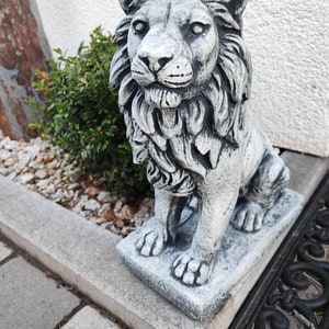 Könnte beinhalten: Eine graue Betonlöwenstatue mit einer detaillierten Mähne, die auf einem quadratischen Sockel sitzt. Die Statue steht im Freien auf einem Steinweg.