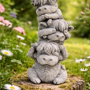 May include: A stacked stone sculpture of three Highland cows, each with shaggy fur and horns, in a garden setting. The sculpture is gray and sits on a tree stump surrounded by flowers and greenery.