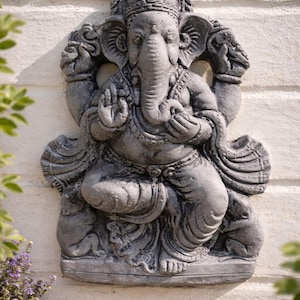 Può includere: Statua in pietra grigia di Ganesha, divinità indù, seduta con le mani in gesto di benedizione. La figura con la testa di elefante è adornata da intagli dettagliati e poggia su una base rettangolare. La statua è montata su un muro di mattoni bianchi.