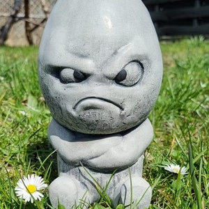 Könnte beinhalten: Ein grauer Steingarten-Zwerg mit einem mürrischen Gesichtsausdruck, der in einem Bett aus grünem Gras mit weißen Gänseblümchen sitzt.