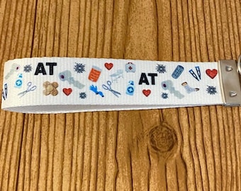 May include: A white fabric wristlet keychain featuring a repeating pattern of medical icons including red hearts, blue scissors, bandages, and pill bottles, with the text AT printed in black. It includes a silver metal clasp and a round key ring.