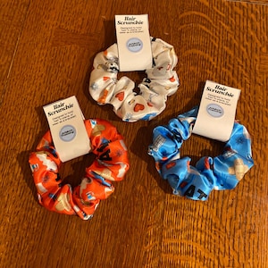May include: Three colorful hair scrunchies with a white background and a variety of designs. The scrunchies are made of satin and have a small tag attached to each one that says "Hair Scrunchie" and "Designed to hold hair in place, or wear as a bracelet!"