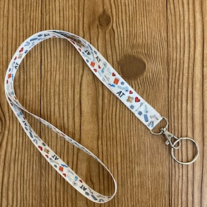 May include: White lanyard with a repeating pattern of hearts, gears, and the letters "AT" in blue and red. The lanyard has a silver-colored metal clasp and key ring attached.