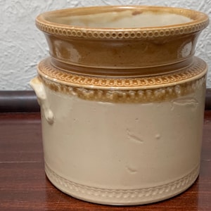 May include: An antique stoneware jar, cylindrical in shape, with a light beige body and brown rim and accents. It has decorative beaded patterns around the top and bottom, and small handles. A classic piece for kitchen or home decor, measuring approximately 15 cm in height.