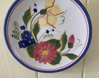 May include: A round ceramic plate with a white base, featuring a hand-painted floral design in shades of yellow, pink, green, and blue. The rim is decorated with a thin pink inner circle and a wider dark blue outer border, framing the central motif.