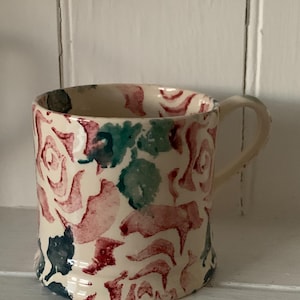 May include: A cream-coloured ceramic mug featuring a hand-painted floral pattern with watercolour-style roses in shades of red, pink, and dark green. The mug has a cylindrical body, a slightly flared base, and a curved handle on the right side.