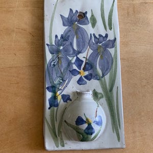 May include: A rectangular white ceramic wall pocket featuring a hand-painted floral design of blue irises with green stems and leaves. The piece includes a small circular opening at the top for hanging and a rounded, bulbous vase section at the bottom.