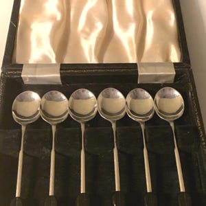 May include: Six silver spoons with rounded bowls and long, thin handles, each with a dark, rounded detail at the end. They are presented in an open black case, lined with cream satin. The spoons are neatly arranged within the case.