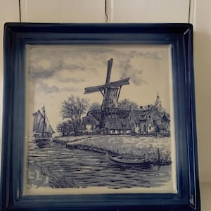 May include: Square ceramic tile featuring a blue and white Dutch windmill scene. The artwork shows a windmill, houses, a sailboat, and a river. The tile has a dark blue border.