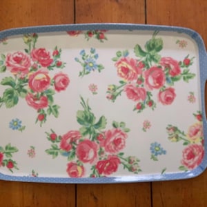 May include: A rectangular serving tray with a white base featuring a floral pattern of pink roses and green leaves. The tray has a light blue border with a white polka dot pattern and two integrated handles on the left and right sides.