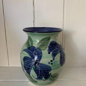 May include: A ceramic vase with a rounded body and flared rim, hand-painted with blue floral designs on a light green background. The rim is dark blue. The vase is approximately 20 cm tall.