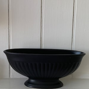 May include: A black decorative bowl with a pedestal base. The bowl has an oval shape and vertical ribbed detailing. It sits on a white shelf against a white paneled wall.