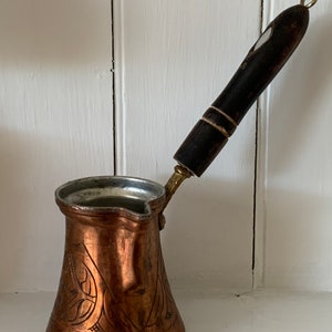 May include: A copper coffee pot with a wide base, a narrow neck, and a pouring spout. The exterior features etched decorative patterns. It has a long, angled handle made of dark brown wood with a small metal loop at the end for hanging.