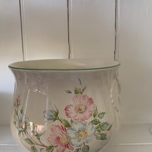 May include: A white ceramic planter with a green rim and floral design. The planter features pink and blue flowers with green leaves.