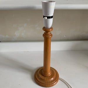 May include: A wooden table lamp with a white bulb socket. The lamp has a light brown, turned wood base and a matching stem. A white electrical cord extends from the base.